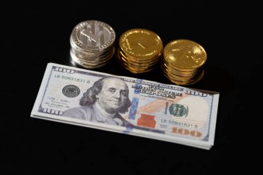 Three distinct stacks of gold and silver-colored crypto coins (Bitcoin, Ethereum, Litecoin) placed behind a pile of $100 USD bills, digital finance with traditional cash assets.