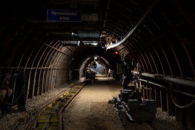 Karanlık, dar bir maden tüneli yerin derinliklerine uzanır, sadece soluk bir yapay ışıkla aydınlatılır. Toz, kaya duvarları ve raylar aktif kömür çıkarmanın yoğun ve ağır atmosferini vurgular.