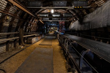 Karanlık, dar bir maden tüneli yerin derinliklerine uzanır, sadece soluk bir yapay ışıkla aydınlatılır. Toz, kaya duvarları ve raylar aktif kömür çıkarmanın yoğun ve ağır atmosferini vurgular.