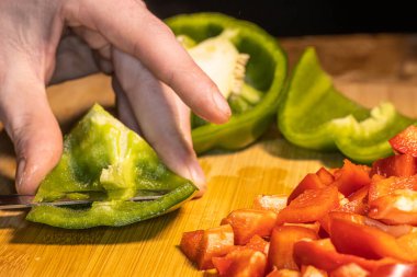 Taze kırmızı, yeşil ve sarı dolma biberler tahta bir kesme tahtasının üzerinde bıçakla doğranıyor. Sahnede sağlıklı yemekler, yemek hazırlama ve renkli sebzeler mutfak ortamında gösteriliyor..