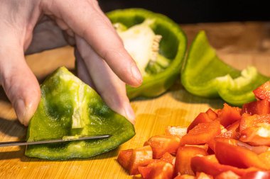 Taze kırmızı, yeşil ve sarı dolma biberler tahta bir kesme tahtasının üzerinde bıçakla doğranıyor. Sahnede sağlıklı yemekler, yemek hazırlama ve renkli sebzeler mutfak ortamında gösteriliyor..