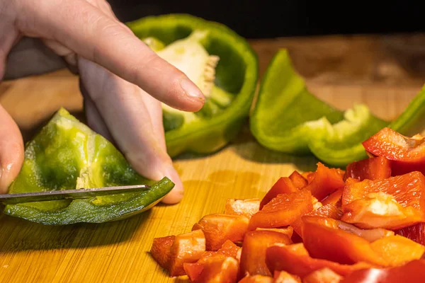 Taze kırmızı, yeşil ve sarı dolma biberler tahta bir kesme tahtasının üzerinde bıçakla doğranıyor. Sahnede sağlıklı yemekler, yemek hazırlama ve renkli sebzeler mutfak ortamında gösteriliyor..