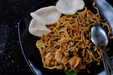 Mie Goreng 'in büyük karideslerle karışık koyu kahverengi soslu lezzetli erişteleri içeren ve siyah bir tabakta iki büyük beyaz karides krakeriyle servis edilen detaylı ve yakından çekilmiş bir fotoğrafı.