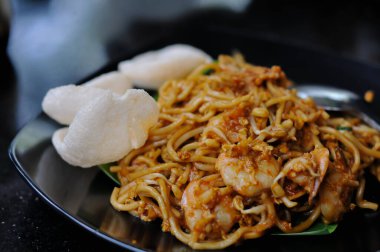 Mie Goreng 'in büyük karideslerle karışık koyu kahverengi soslu lezzetli erişteleri içeren ve siyah bir tabakta iki büyük beyaz karides krakeriyle servis edilen detaylı ve yakından çekilmiş bir fotoğrafı.