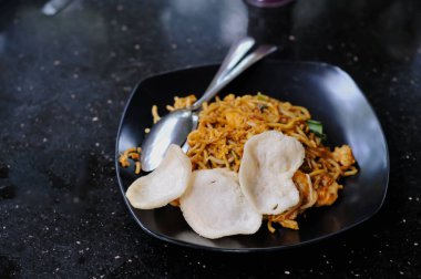 Mie Goreng 'in büyük karideslerle karışık koyu kahverengi soslu lezzetli erişteleri içeren ve siyah bir tabakta iki büyük beyaz karides krakeriyle servis edilen detaylı ve yakından çekilmiş bir fotoğrafı.