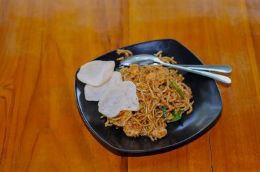 Siyah bir tabakta beyaz kraker ve sıcak ahşap bir masaya yerleştirilmiş Endonezyalı Mie Goreng 'in (kızarmış erişteler) servis edişinin genel manzarası..