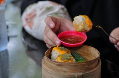 Bir el, yuvarlak, portakallı bir Siomay (gamsum) hamur köftesini kaldırmak için çatal tutuyor, bambu buharlı buharlı tencerenin üstündeki kırmızı bir biber sosuna batırıyor, Asya yemek kültürünü ve Çin mutfağını ön plana çıkarıyor.,