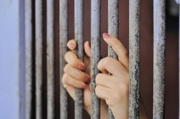  Пара мужских рук и лоб прижаты к жестким белым металлическим решеткам тюремной камеры, с лицом, затемненным глубокой тенью, передающими темы заточения, плена, вины и ЭМ