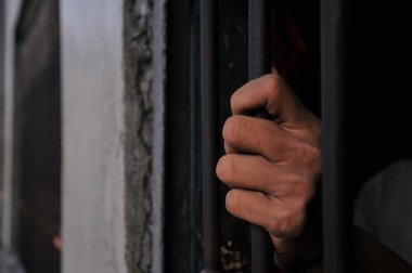 Mahkûmun elleri siyah arka planda parmaklıklar ardında.