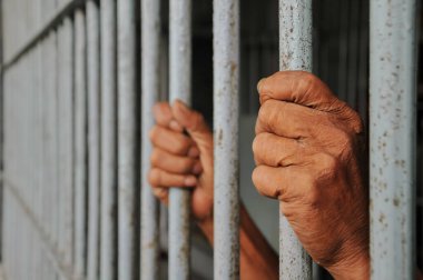 Mahkûmun elleri siyah arka planda parmaklıklar ardında.