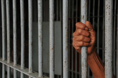 Mahkûmun elleri siyah arka planda parmaklıklar ardında.