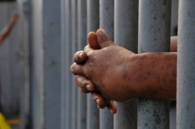 Mahkûmun elleri siyah arka planda parmaklıklar ardında.