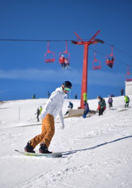 Mavi gökyüzü ve arka planda telesiyej ile yokuş aşağı snowboard üzerinde genç adam