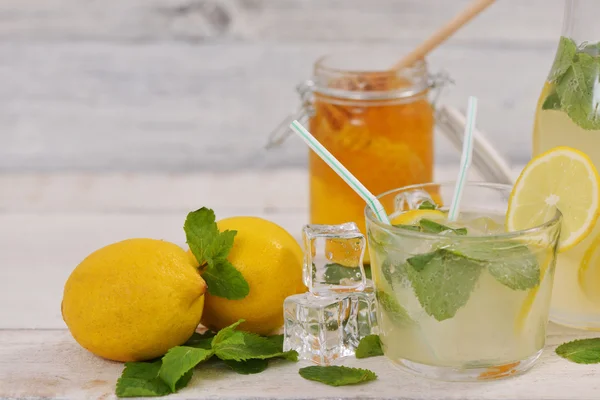 Glass of lemonade with lemon slice, mint leaves and ice cubes near lemons, lemonade bottle and honey jar on rustic white wood — Stock Photo, Image