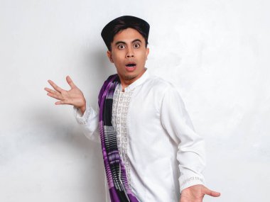Yetişkin Endonezyalı Müslüman adam şok olmuş ve komik bir yüz ifadesiyle stüdyoda beyaz arka planda bekliyor.