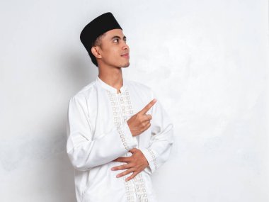 Gülümseyen Endonezyalı Müslüman adam midesini tutuyor ve beyaz arka plandaki fotokopi alanını işaret ediyor, aç hissediyor veya Ramazan ayında oruç tutma kavramı sırasında yemek öneriyor.