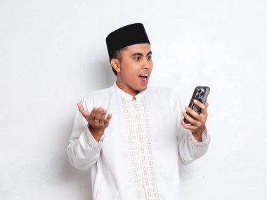 Yetişkin Endonezyalı Müslüman bir adam elinde tutuyor ve beyaz arka planda stüdyoda şaşırmış bir ifadeyle internetten uyarı, mesaj veya Ramazan dijital sunuyor.