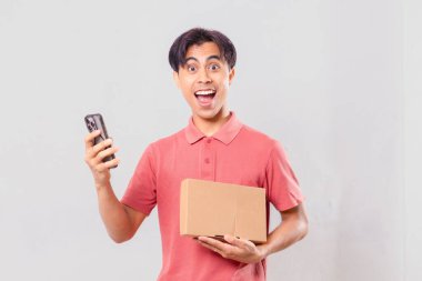 Karton paket kutusunu dış ticaret alışveriş konsepti için sade arka planda tutarken cep telefonu kullanan Asyalı bir adam şaşırdı..
