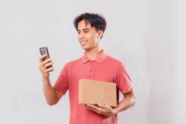 Gülümseyen Asyalı adam elinde karton paket kutusu tutarken cep telefonu kullanıyor. Ekommerce teslimat kavramı için arka planda izole edilmiş..