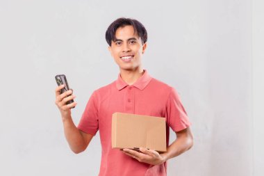 Gülümseyen Asyalı adam elinde karton paket kutusu tutarken cep telefonu kullanıyor. Ekommerce teslimat kavramı için arka planda izole edilmiş..