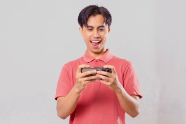 Akıllı telefondan mobil oyun oynayan genç Asyalı adam heyecanlı bir zafer jesti sergiliyor, oyun ve teknoloji reklamcılığı için sade bir arka planda izole edilmiş..