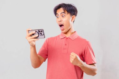 Akıllı telefondan mobil oyun oynayan genç Asyalı adam heyecanlı bir zafer jesti sergiliyor, oyun ve teknoloji reklamcılığı için sade bir arka planda izole edilmiş..