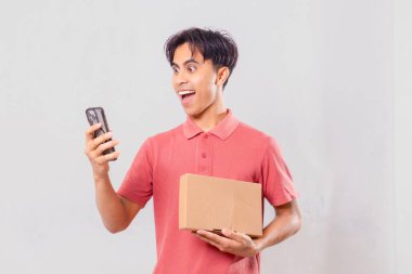 Karton paket kutusunu dış ticaret alışveriş konsepti için sade arka planda tutarken cep telefonu kullanan Asyalı bir adam şaşırdı..