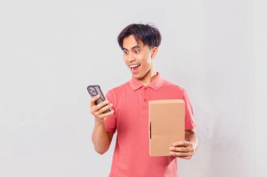 Karton paket kutusunu dış ticaret alışveriş konsepti için sade arka planda tutarken cep telefonu kullanan Asyalı bir adam şaşırdı..