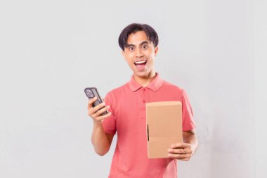 Karton paket kutusunu dış ticaret alışveriş konsepti için sade arka planda tutarken cep telefonu kullanan Asyalı bir adam şaşırdı..