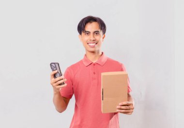 Gülümseyen Asyalı adam elinde karton paket kutusu tutarken cep telefonu kullanıyor. Ekommerce teslimat kavramı için arka planda izole edilmiş..