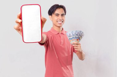 Neşeli Asyalı genç adam elinde para tutuyor ve boş akıllı telefon ekranı gösteriyor. Dijital ödeme ve finans kavramı için sade arka planda izole edilmiş..