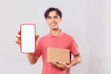 Neşeli, genç, yetişkin Asyalı adam boş akıllı telefon ekran maketi gösteriyor ve paket kutusunu teslimat servisi tanıtımı için sade bir arka planda tutuyor..