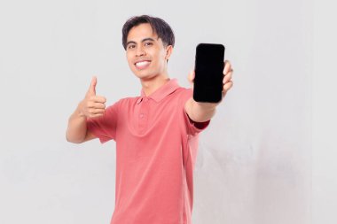 Neşeli Asyalı genç adam boş bir cep telefonu ekranı gösteriyor. Başparmak hareketi yapıyor.