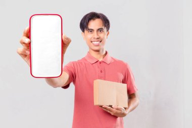 Neşeli, genç, yetişkin Asyalı adam boş akıllı telefon ekran maketi gösteriyor ve paket kutusunu teslimat servisi tanıtımı için sade bir arka planda tutuyor..