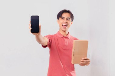 Heyecanlı Asyalı adam boş cep telefonu ekranı gösterirken, karton paket kutusunu dış ticaret kavramı için izole edilmiş olarak tutuyor..
