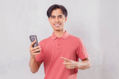 Arka planda izole edilmiş akıllı telefonu işaret eden mutlu Asyalı adam.