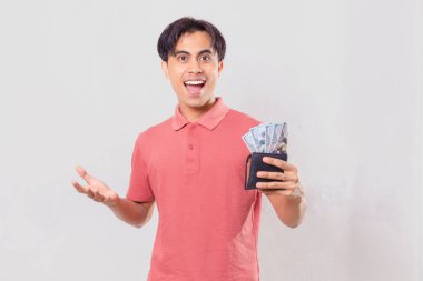 Şaşırmış Güneydoğu Asyalı genç adam elinde bir cüzdan dolusu nakit para tutarken, satış promosyonu, mali sonuçlar, gelir mesajları ve ticari reklamlar için kullanılıyor..