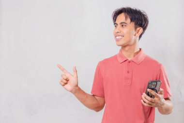 Neşeli genç Asyalı adam cep telefonu tutarken yan tarafı işaret ediyor, arka planda izole edilmiş, reklam, pazarlama ve sosyal medya kullanımı için mükemmel.