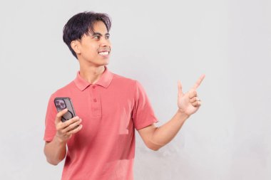 Neşeli genç Asyalı adam cep telefonu tutarken yan tarafı işaret ediyor, arka planda izole edilmiş, reklam, pazarlama ve sosyal medya kullanımı için mükemmel.