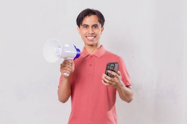 Elinde megafonla gülümseyen genç Asyalı adam, cep telefonu kullanırken kameraya bakıyor, iletişim ve pazarlamayı gösteriyor..