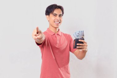 Neşeli Güneydoğu Asyalı genç adam elinde para dolu bir cüzdan tutuyor ve ileri doğru işaret ediyor, satış promosyonu, özel teklifler, ödüller ve ticari reklam kullanımı için teşvik ediyor.