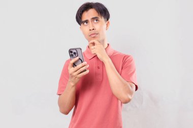 Ciddi bir Asyalı genç adam cep telefonu tutuyor ve kullanıyor. Düşünen bir ifadeyle bakıyor. İş ve teknoloji için sade bir arka planda izole edilmiş..