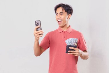 Cep telefonuna bakarken içi para dolu bir cüzdan tutan heyecanlı Asyalı genç adam, beyaz arka planda tanıtım, satış, pazarlama ve ticari reklam kullanımı için izole edilmiş.