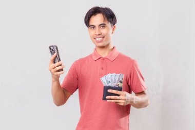 Akıllı telefon kullanırken içi para dolu bir cüzdan tutan kendine güvenen Güneydoğu Asyalı genç adam, beyaz arka planda ticari promosyon, pazarlama, finans ve reklam kullanımı için izole edilmiş..