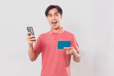 Kameraya bakarken banka hesap cüzdanı tutan genç adam şaşırdı. Stüdyoda beyaz bir arka planda izole edilmiş.