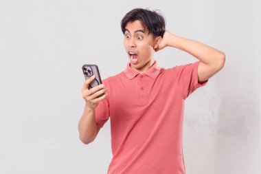 Şok olmuş genç Asyalı adam cep telefonu kullanıyor, arka planda izole edilmiş.