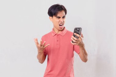 Genç Asyalı adam öfkeyle cep telefonunu kullanıyor, sade arka planda izole edilmiş, ticari ve editörel kullanım için hayal kırıklığı ve duygu gösteriyor.