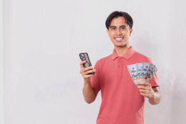 Cep telefonu kullanırken elinde nakit parayla gülümseyen genç bir adam. Stüdyoda beyaz bir arka planda izole edilmiş.