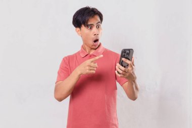 Genç Asyalı adam öfkeyle cep telefonunu kullanıyor, sade arka planda izole edilmiş, ticari ve editörel kullanım için hayal kırıklığı ve duygu gösteriyor.