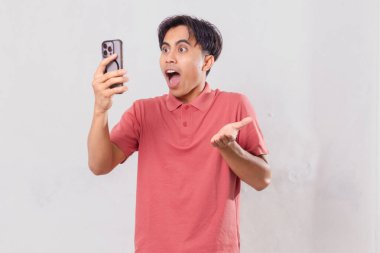 Yetişkin Güneydoğu Asyalı bir adam beyaz arka planda izole edilmiş, şok olmuş bir ifadeyle cep telefonuna bakıyor.